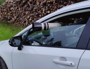Auto on parkkeerattu pellon laitaan puupinon viereen. Kuljettajan ikkunan raosta työntyy kameran teleobjektiivi.